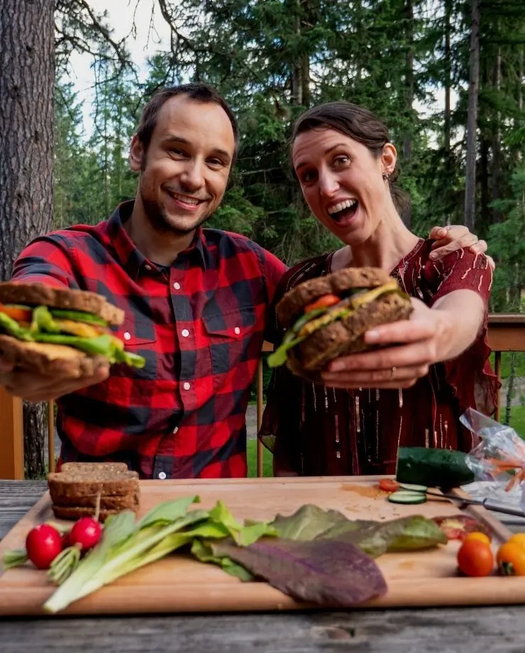 Two people smiling outdoors as they hold out sandwiches toward the camera, with fresh vegetables on a cutting board
