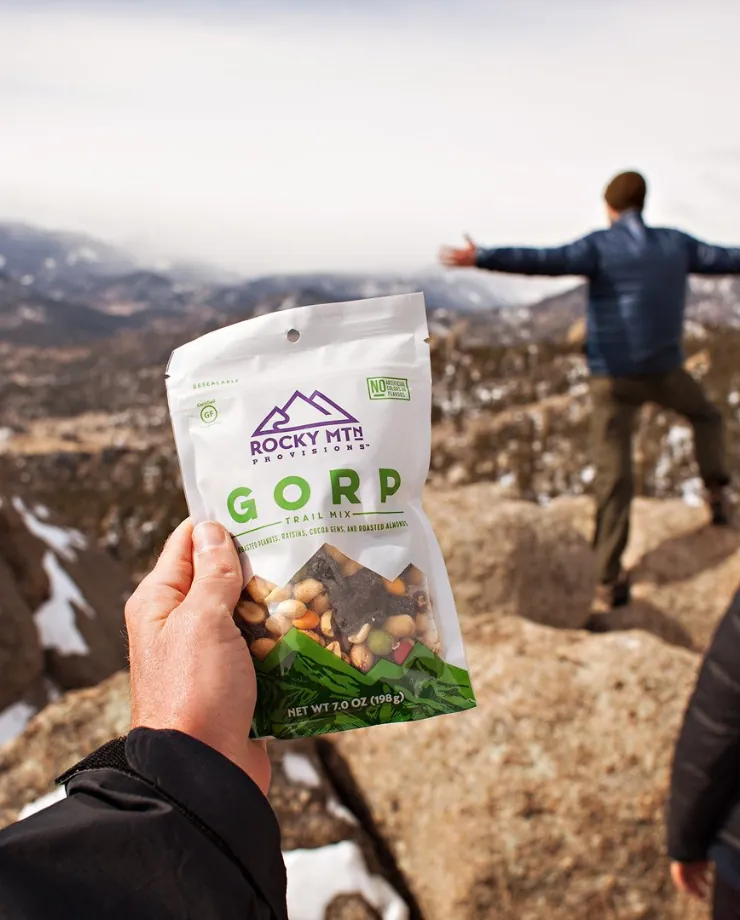 Hand holding a bag of Rocky Mtn GORP trail mix on a mountaintop as a hiker stretches in the background